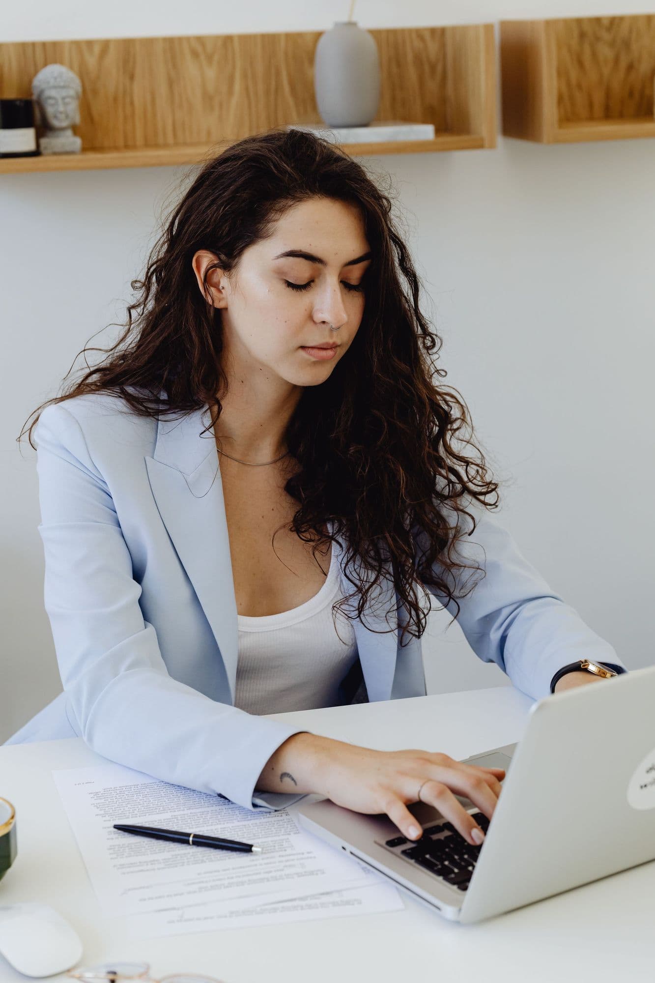 A woman in a pale blue blazer types at her laptop beside a printed draft and a pen.