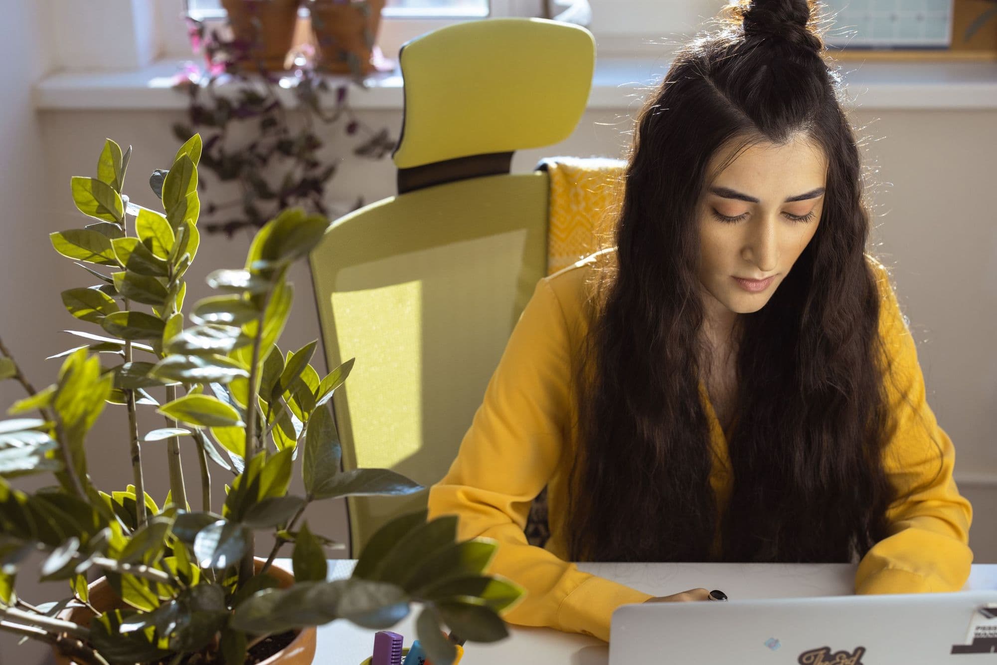 A woman in a yellow sweater works through a task on her laptop at a desk in a sunny room.