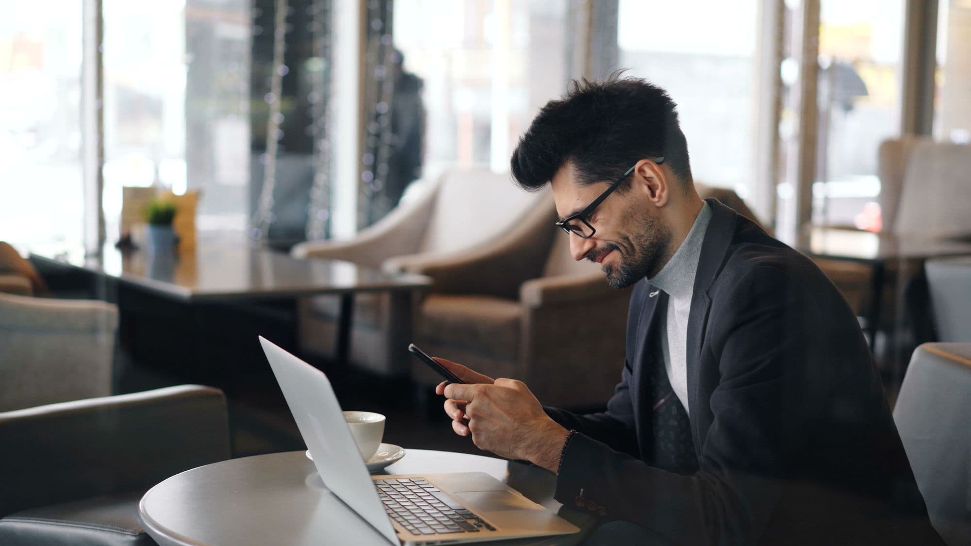 A man in a blazer smiles at a message on his phone at a café table, his laptop open beside him.