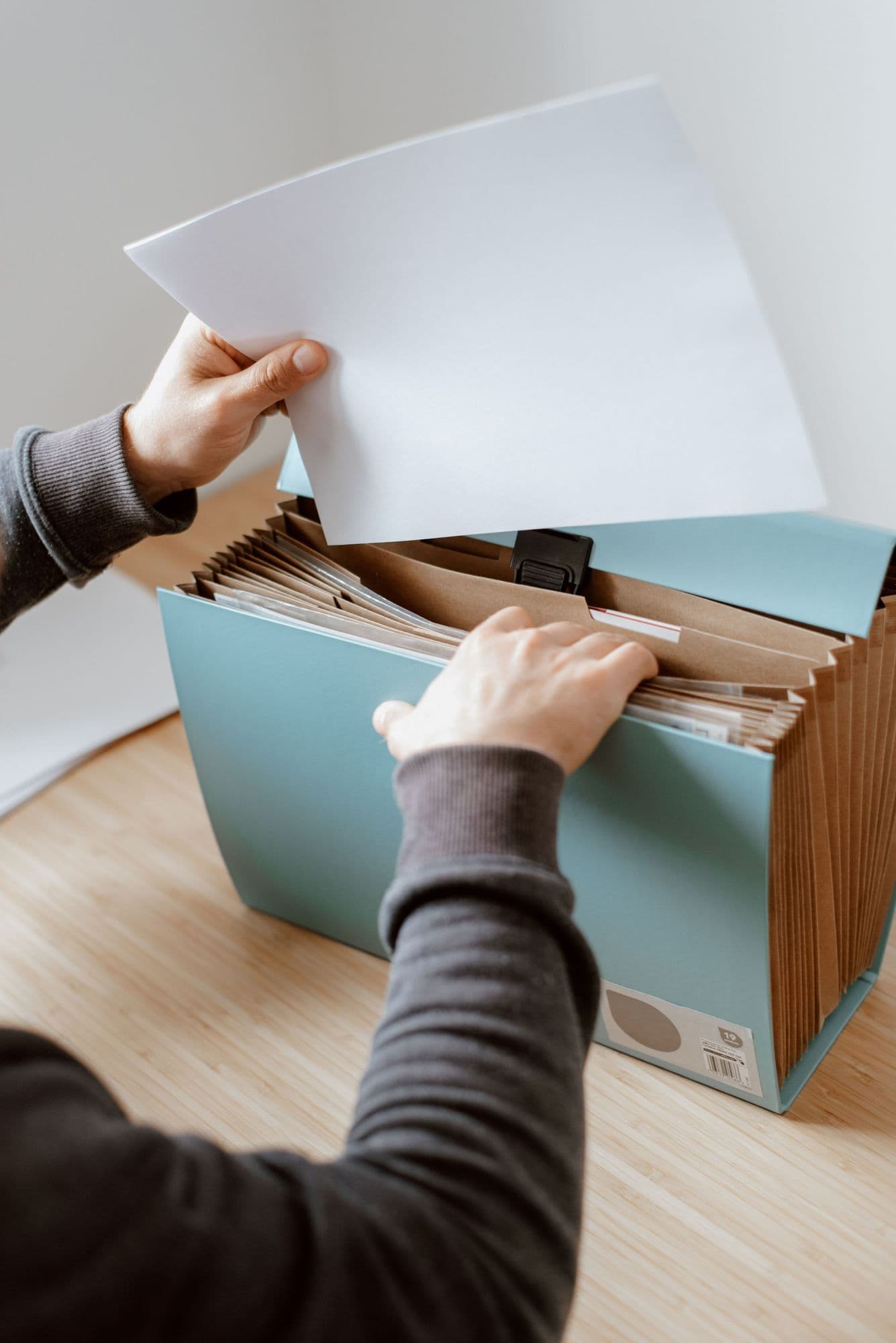 Hands file a sheet of paper into an accordion folder on a wooden desk.